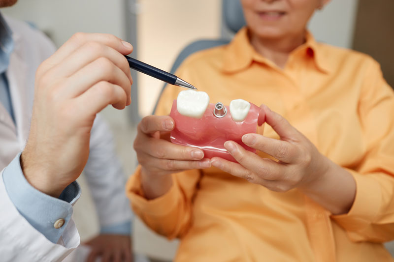 Closeup of unrecognizable dentist pointing at tooth model while explaining dental implant surgery to patient in clinic, discussing the dental implant treatment process